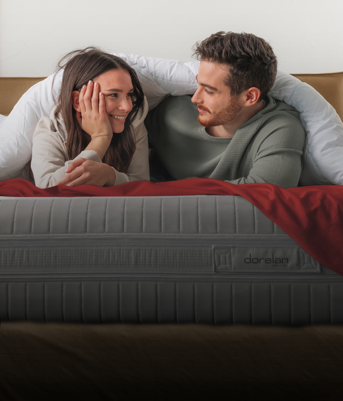 Una coppia distesa su un letto Dorelan, avvolta in una soffice coperta bianca e immersa in un’atmosfera intima e accogliente. Il materasso è in primo piano, simbolo di comfort e qualità, mentre il dettaglio della coperta rossa richiama il calore delle festività.
